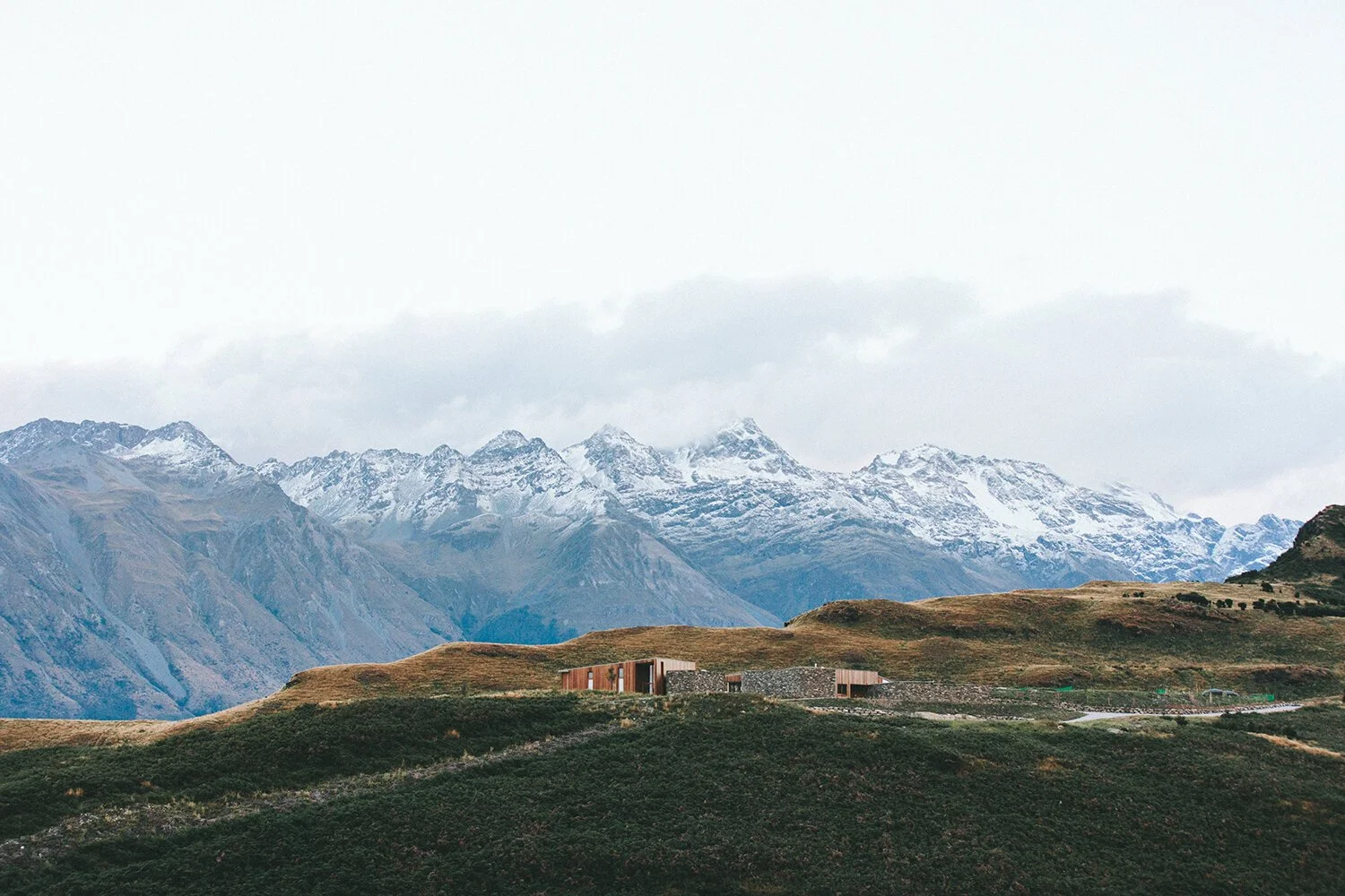 A landscape view of mountains with snow on their peaks, a cloudy sky, and a grassy hillside with a modern building.